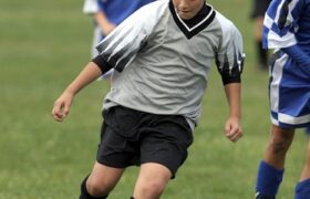 Male soccer player ADK carries the ball down field past opposition.  Young players, full figure shot.  Gray/black uniform.